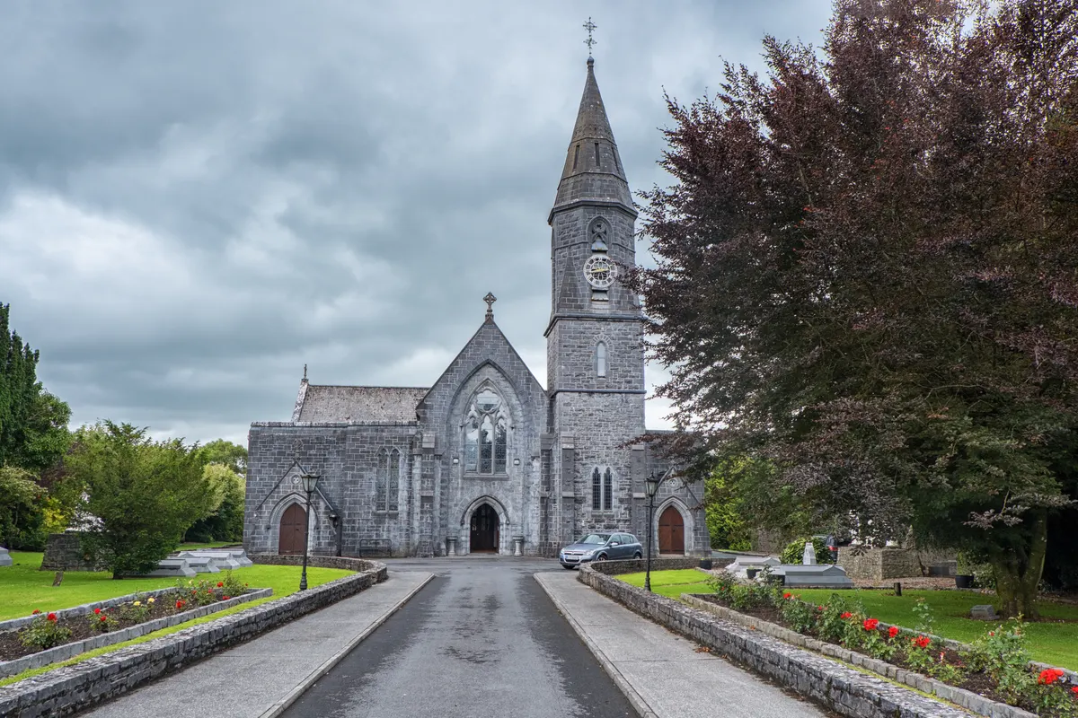 St Mary's Church in Ballinrobe, Co. Mayo, a grey stone Gothic church with a tall spire at the end of a tree-lined drive
